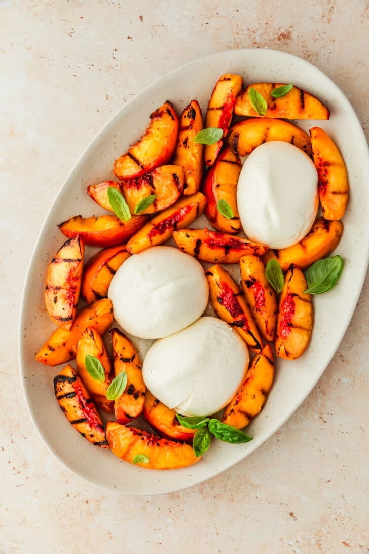 A white platter of grilled peaches, burrata, and basil.