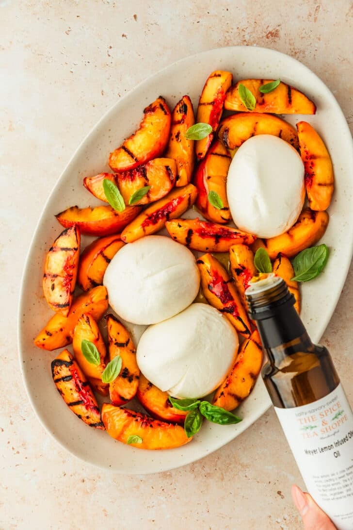 A hand drizzling lemon oil over a white platter of peaches, burrata, and basil.