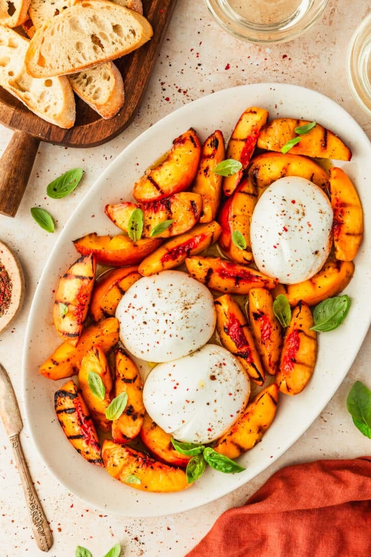 Grilled peaches and burrata appetizer on a white platter next to an orange linen, glasses of white wine, and a wood board of crostini.