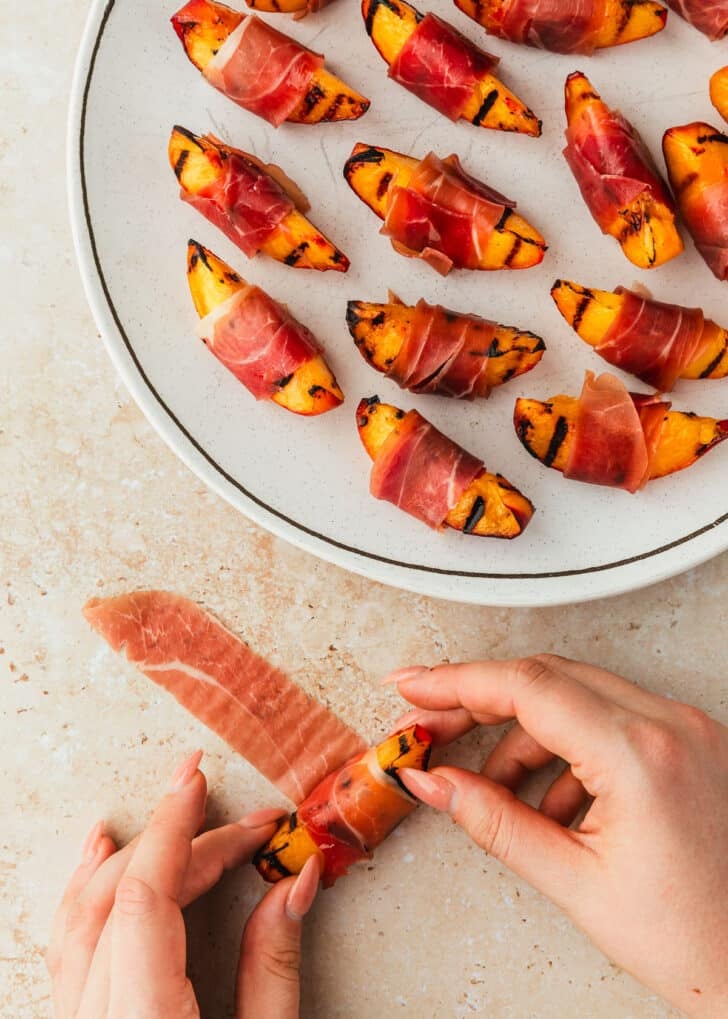 Hands rolling peaches in prosciutto for prosciutto-wrapped peach skewers.