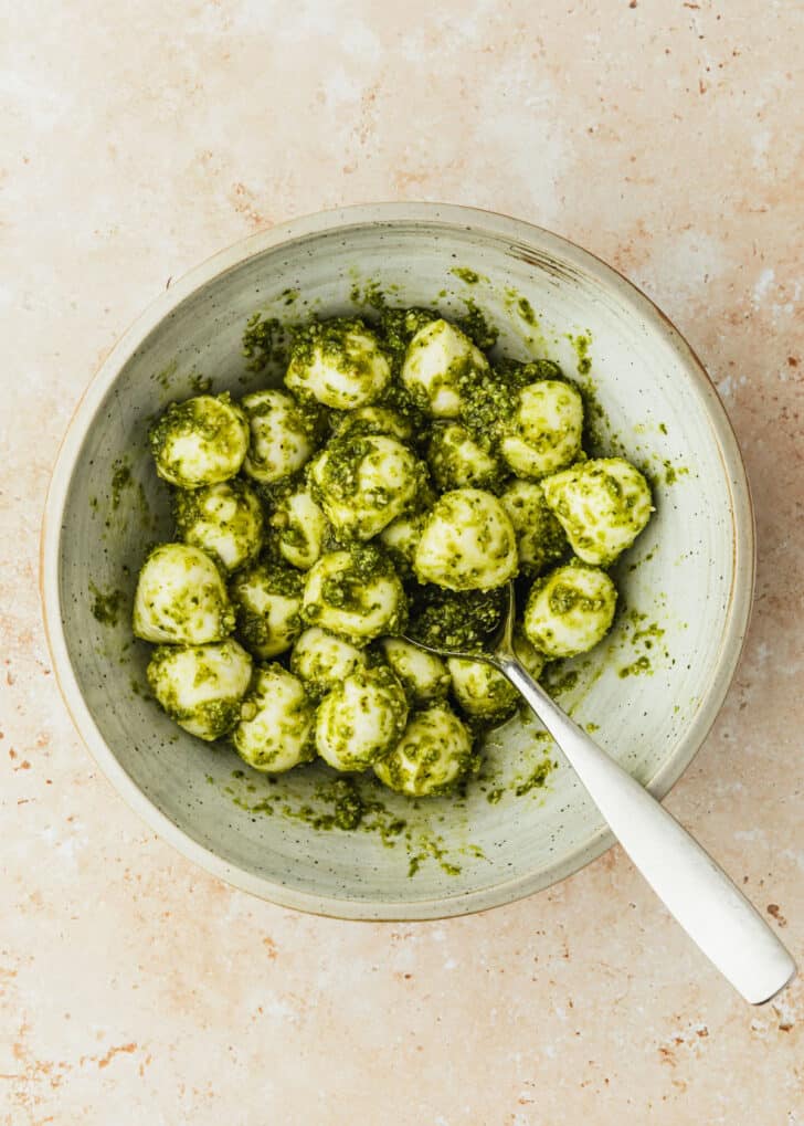 A grey bowl of mozzarella balls covered in pesto on a brown counter.