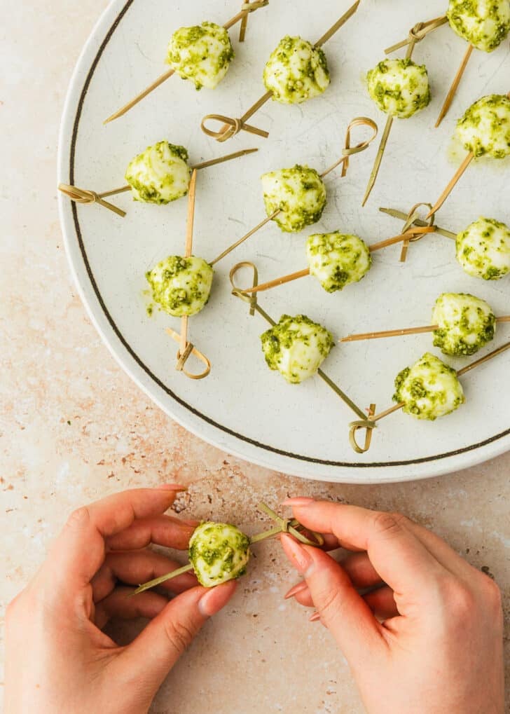 Hands skewering mozzarella balls with pesto for prosciutto-wrapped peach skewers.