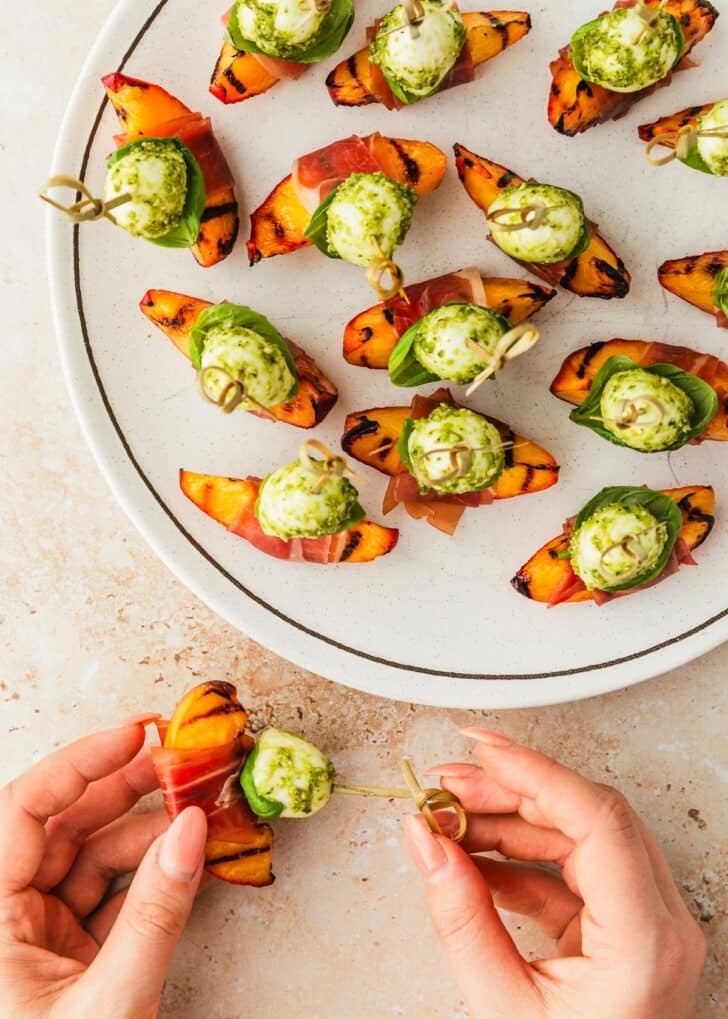 Hands assembling prosciutto-wrapped peach skewers on a brown counter.