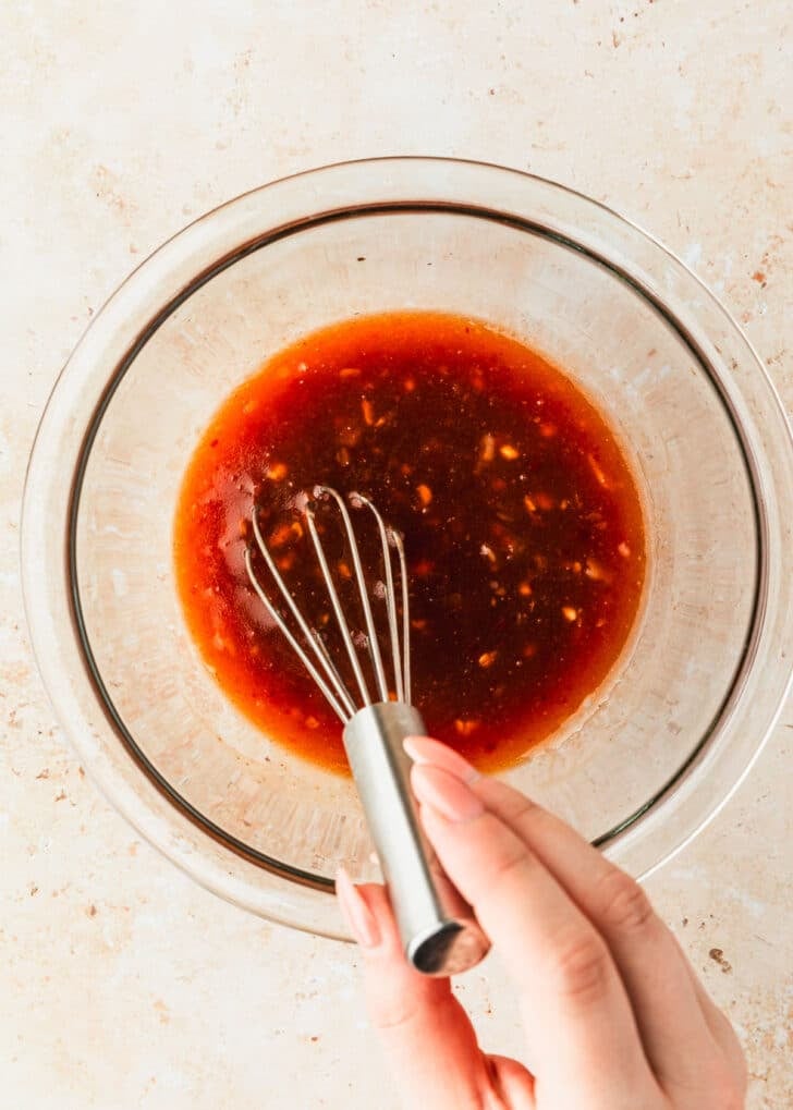 A hand using a whisk to mix sweet chili sauce in a glass bowl.