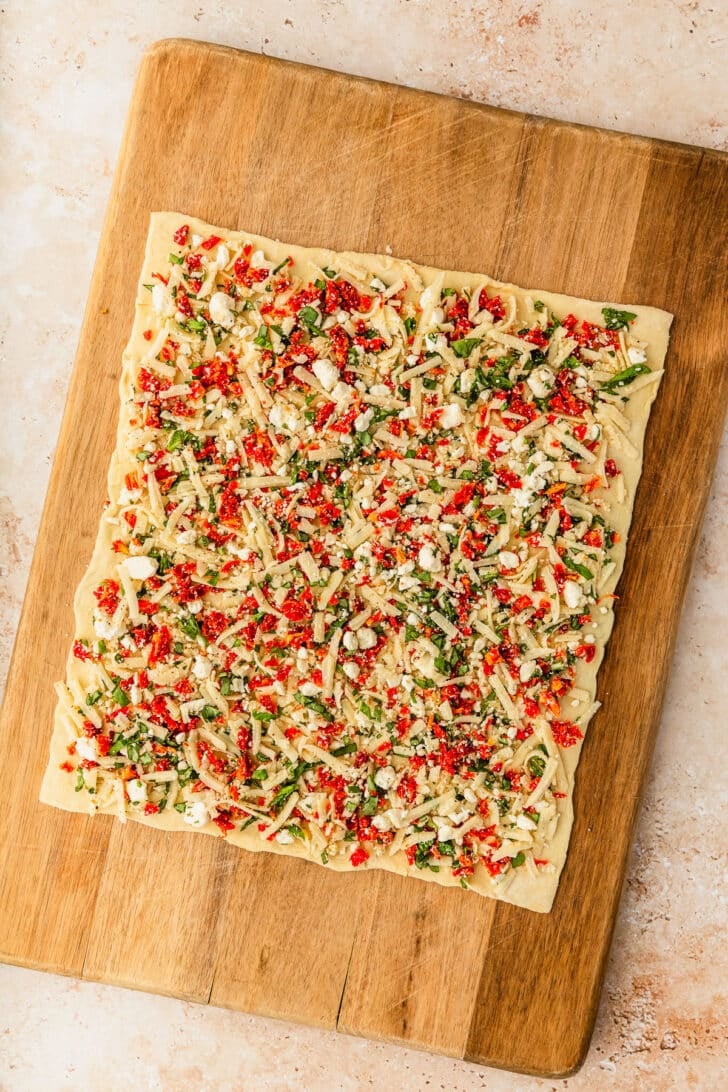 A sheet of puff pastry topped with a cheese tomato mixture on a wood board.