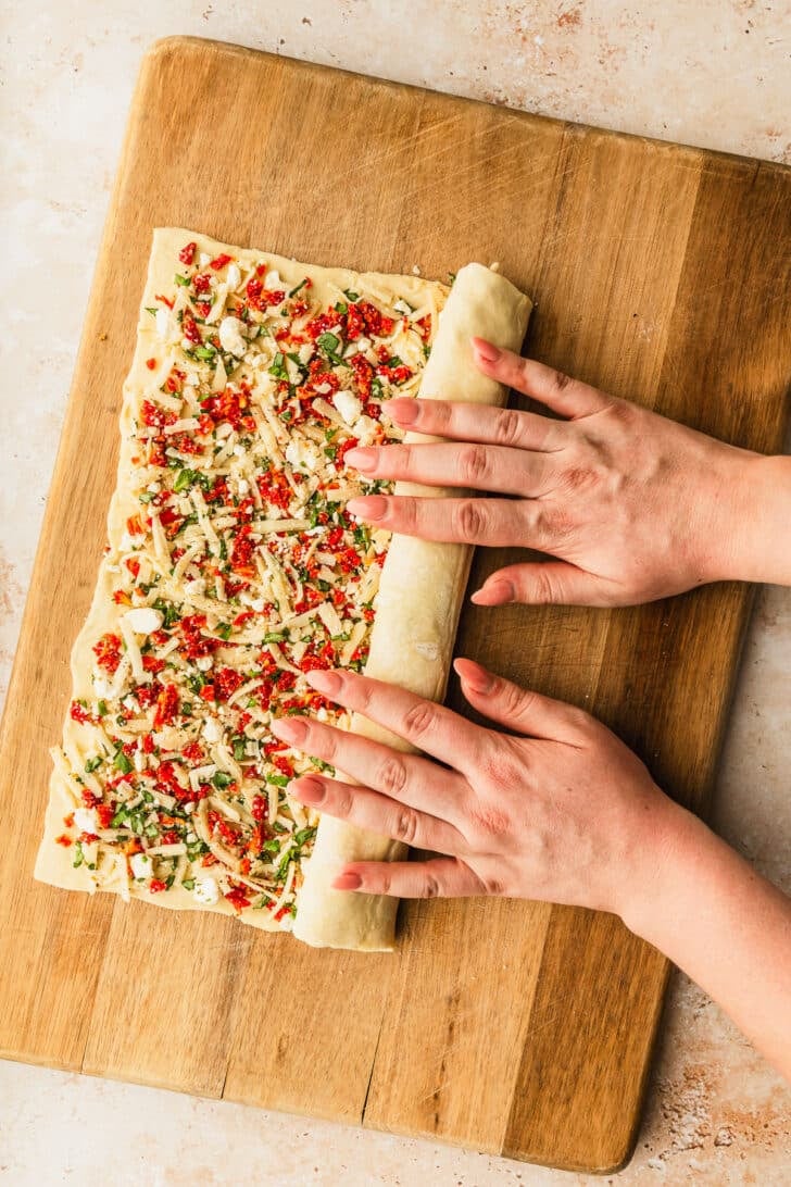 Hands rolling puff pastry into a log on a wood board.