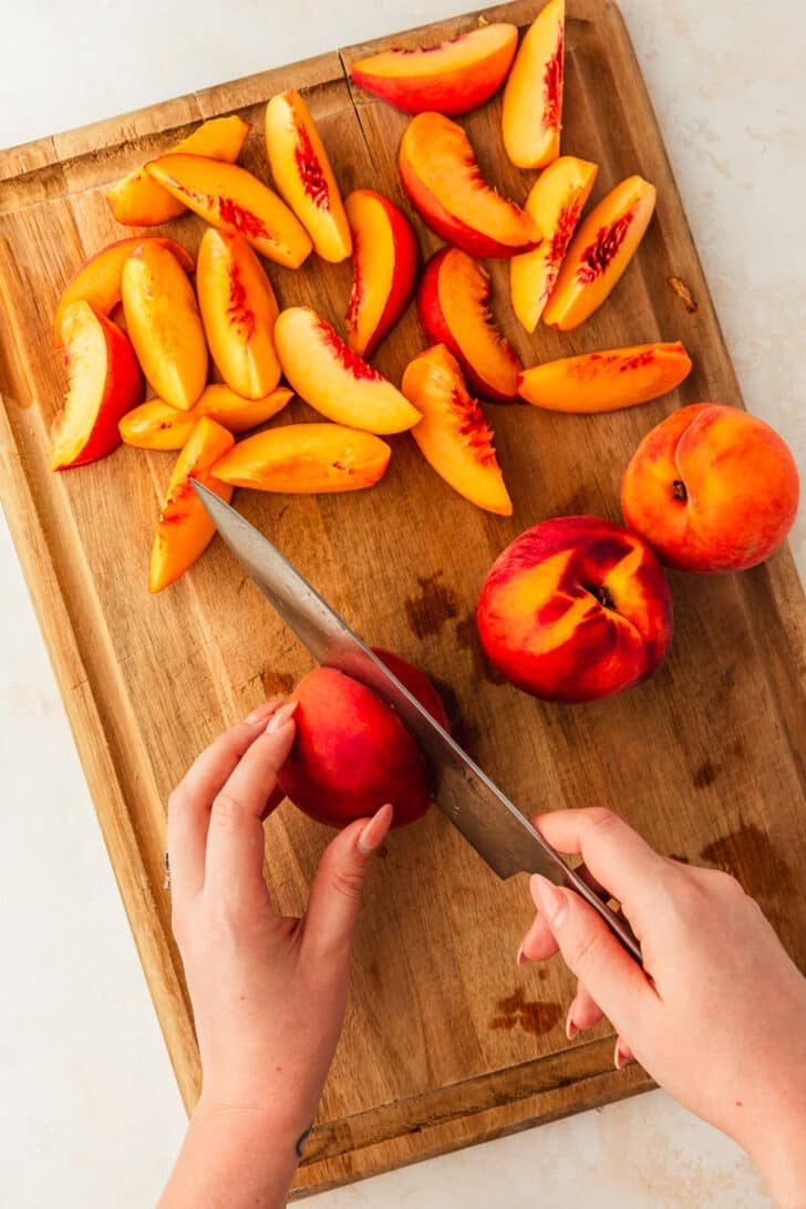 A hand using a knife to slice a peach in half on a wood board of slice peaches.
