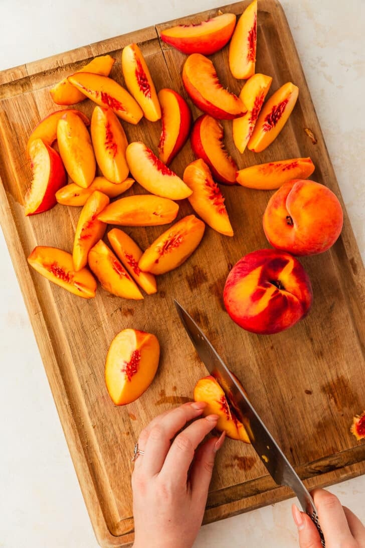 Hands using a knife to slice peaches on a wood board for a peaches and burrata appetizer.