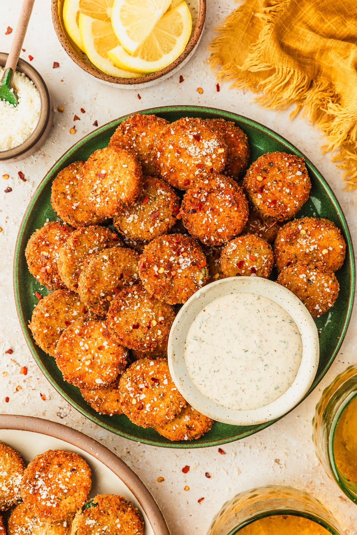 A green plate of fried zucchini and white bowl of ranch on a tan counter next to glasses of beer, a yellow linen, a white bowl of lemon slices, and a brown bowl of parmesan.
