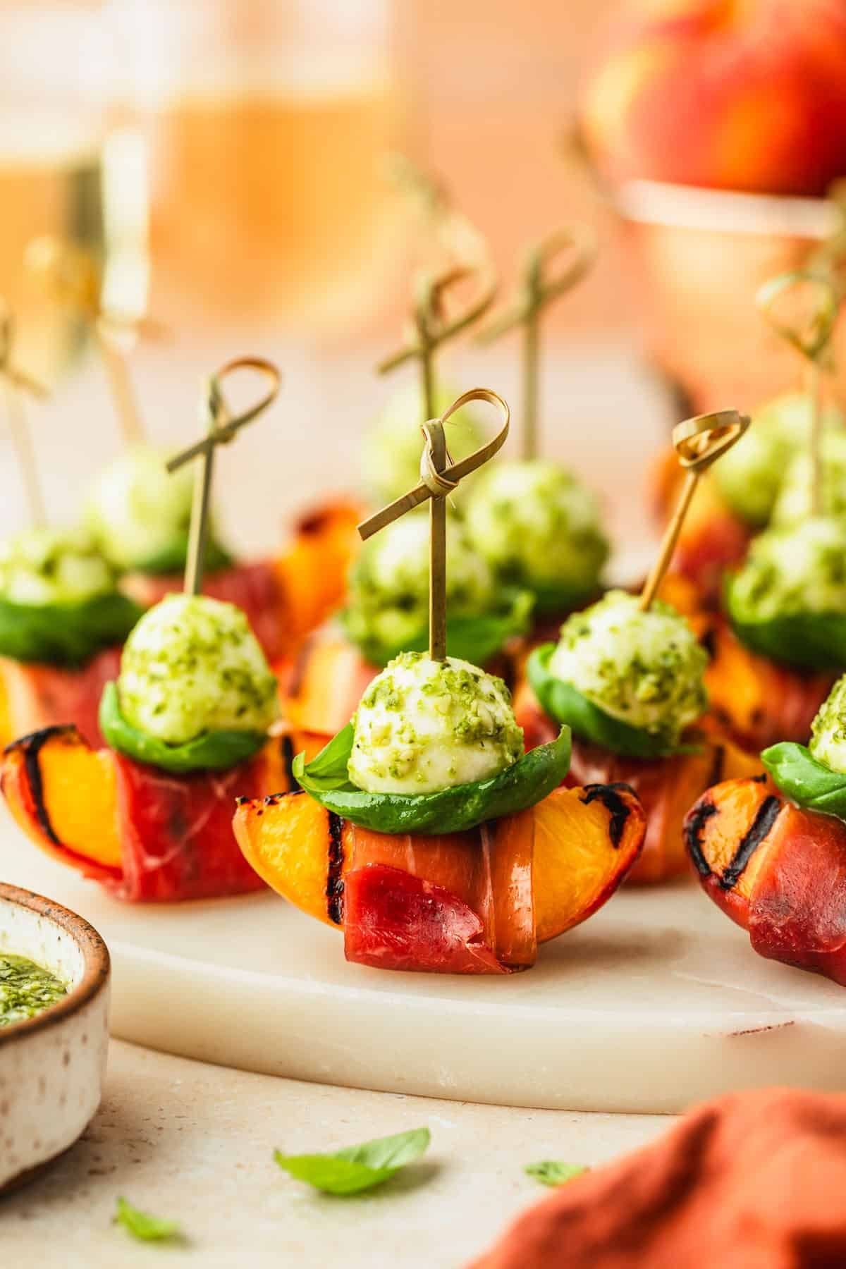 A white platter of prosciutto-wrapped peach skewers on a tan counter next to a white bowl of pesto, orange linen, and copper bowl of peaches.