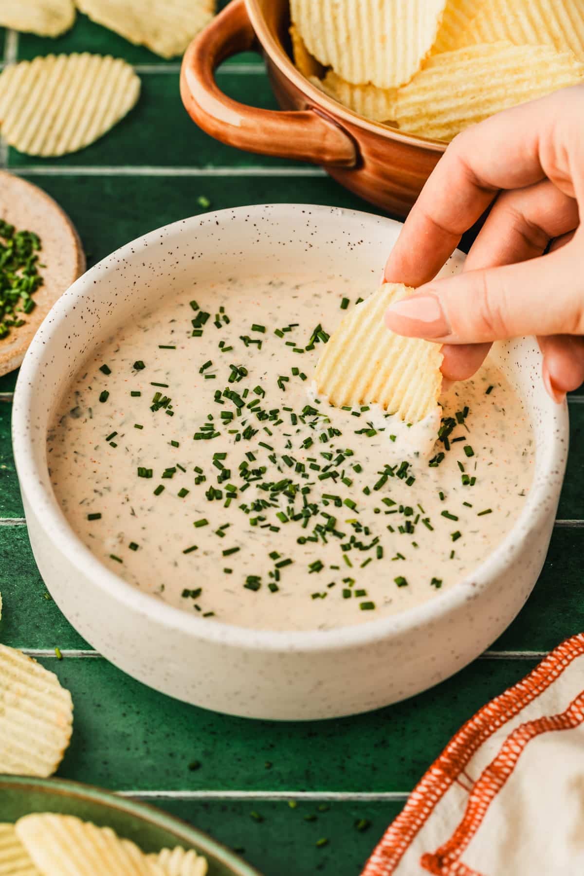 A hand dipping a chip into a white bowl of buttermilk ranch dip on a green tiled counter next to a brown bowl of chips, an orange and white striped linen, and a brown bowl of chives.