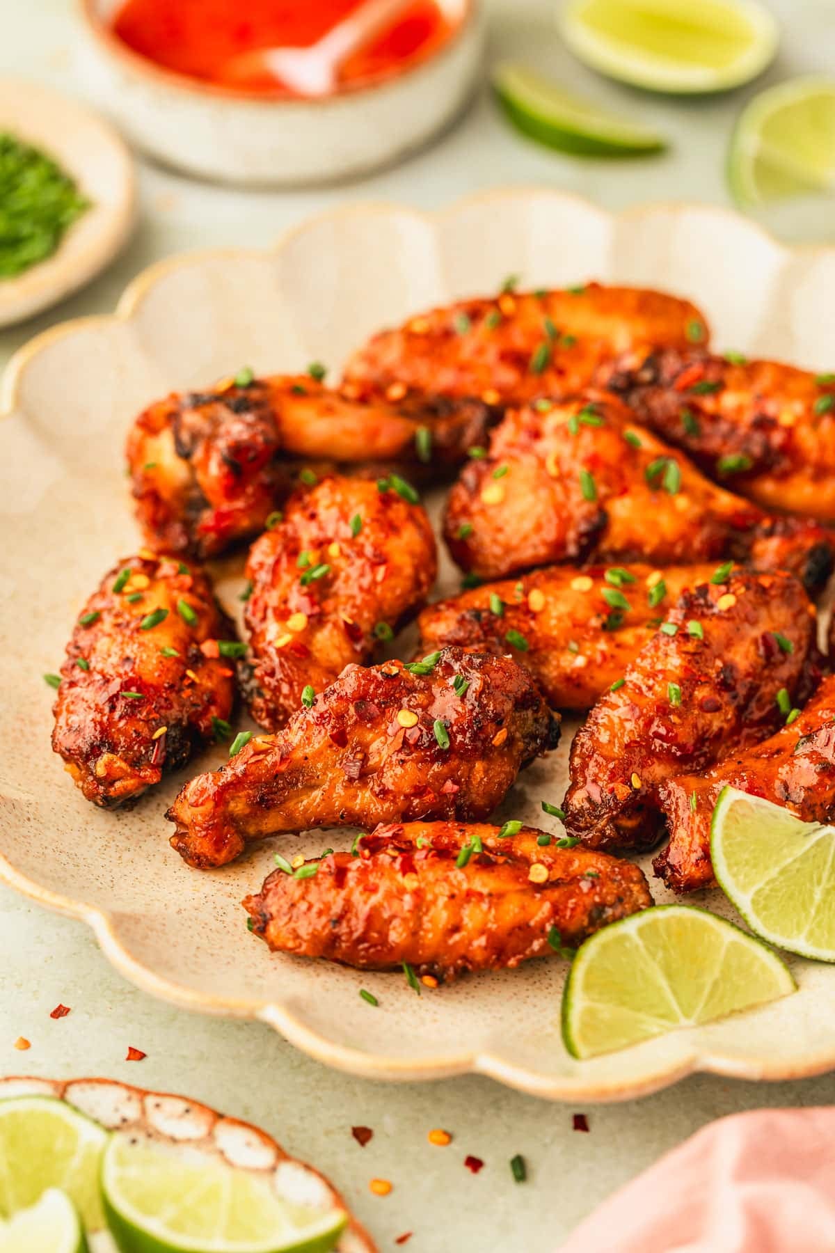 A tan platter of sweet chili chicken wings on a green counter next to a pink linen and brown bowls of limes, chives, and chili sauce.