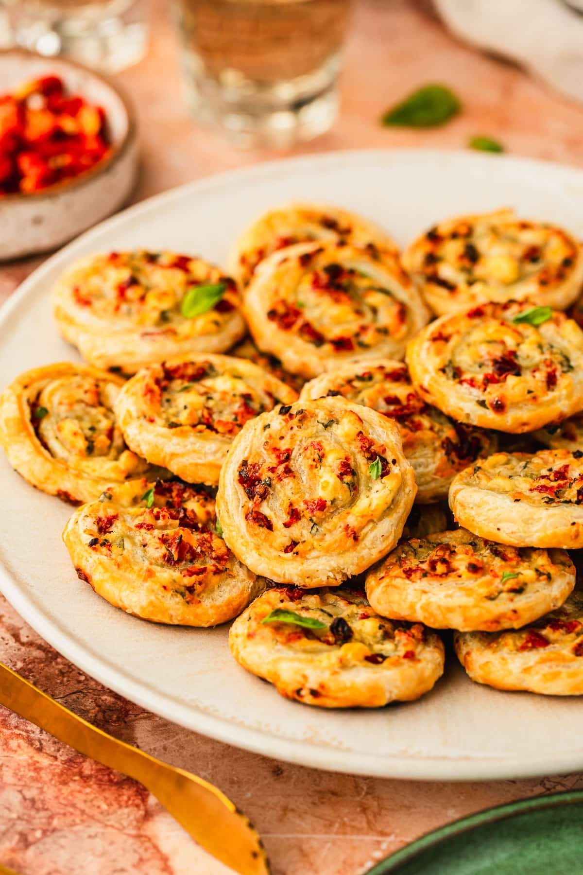 A white platter of sun-dried tomato and cheese pinwheels on an orange marble counter next to a green plate, glasses of white wine, and a white bowl of sun-dried tomatoes.