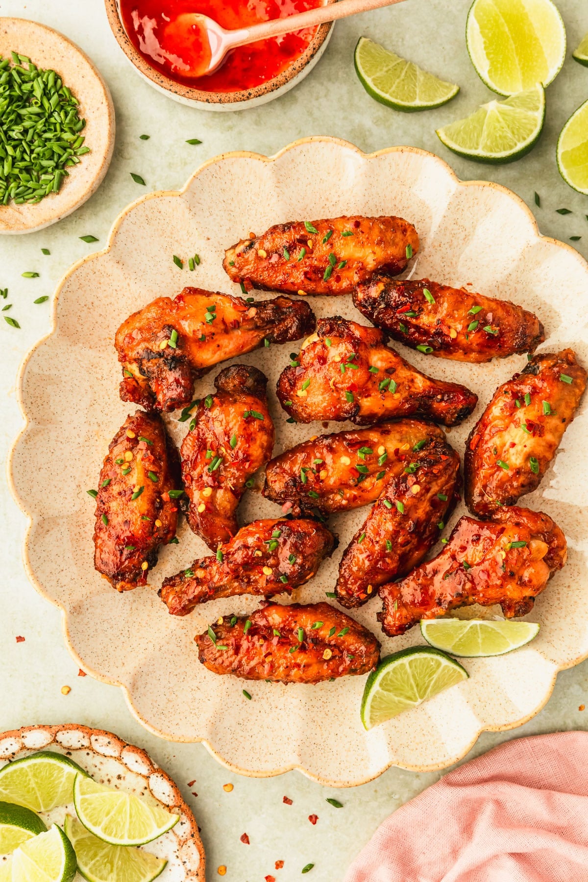 A tan platter of sweet chili chicken wings on a green counter next to brown bowls of limes, chives, and sweet chili sauce.