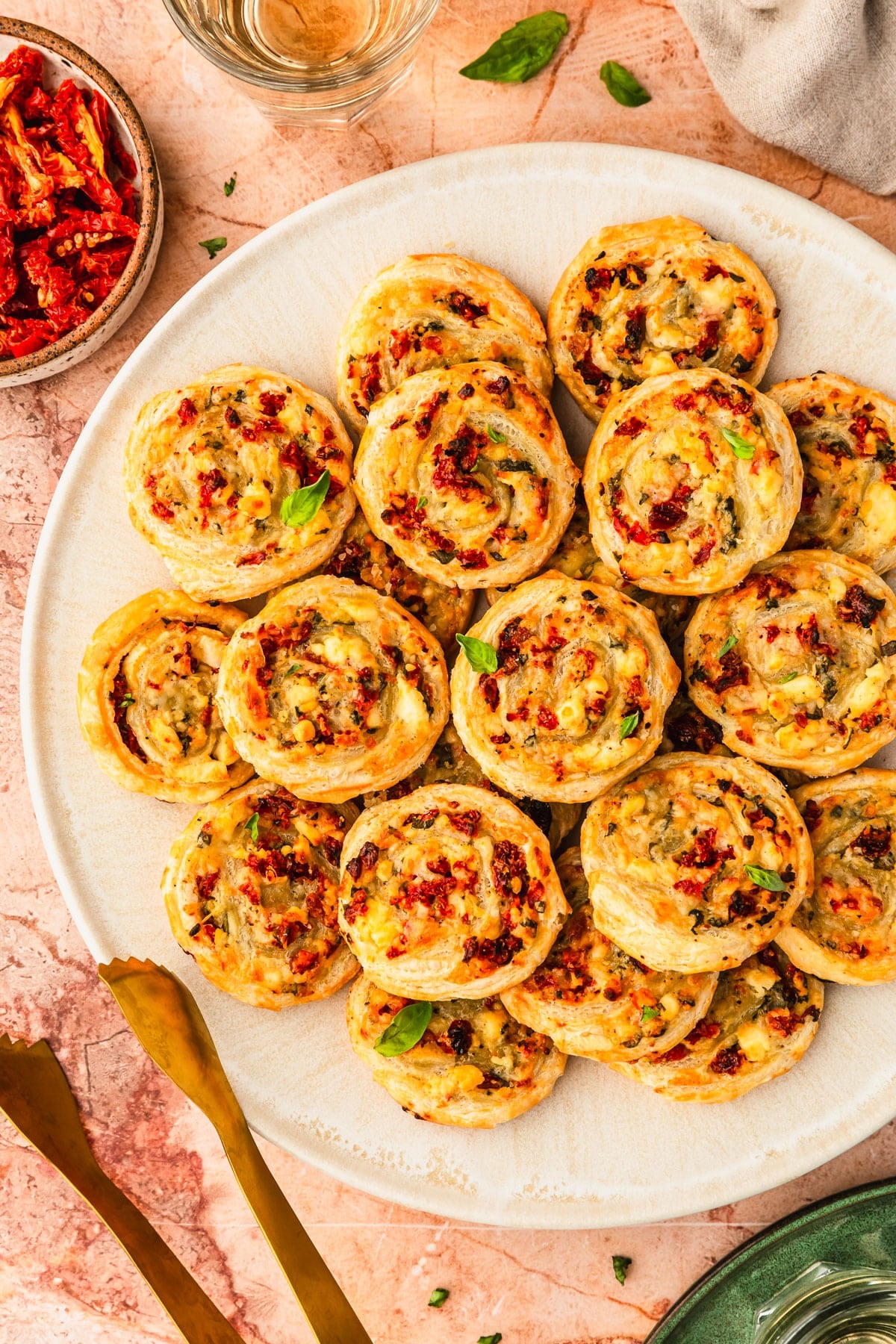 A white platter of sun-dried tomato and cheese pinwheels on an orange marble counter next to glasses of white wine, a tan linen, a green plate, and a white bowl of sun-dried tomatoes.