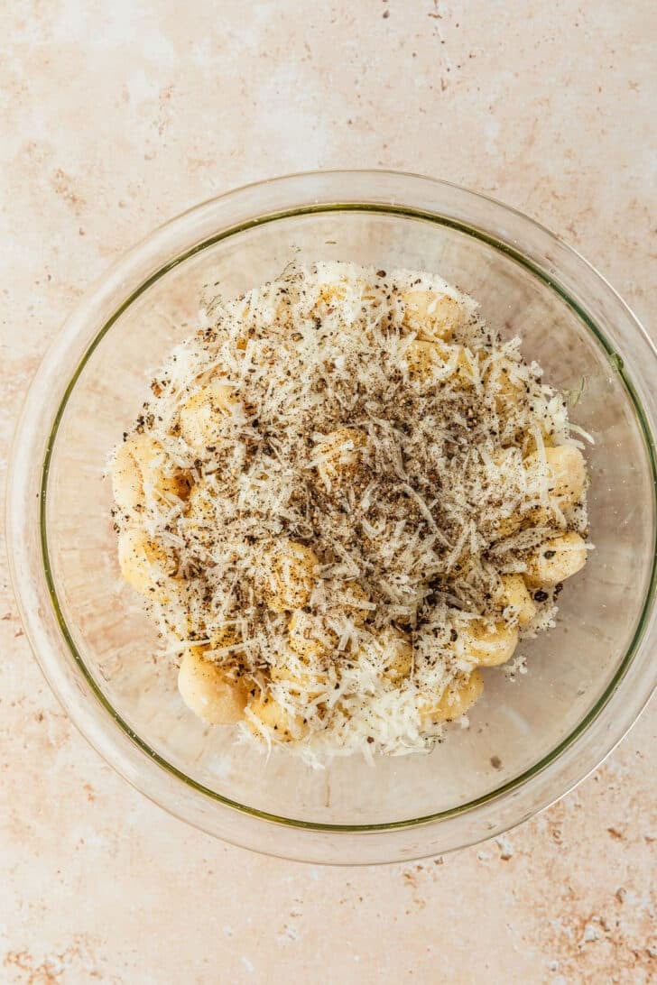 A clear bowl of pasta, parmesan, and black pepper on a tan counter.