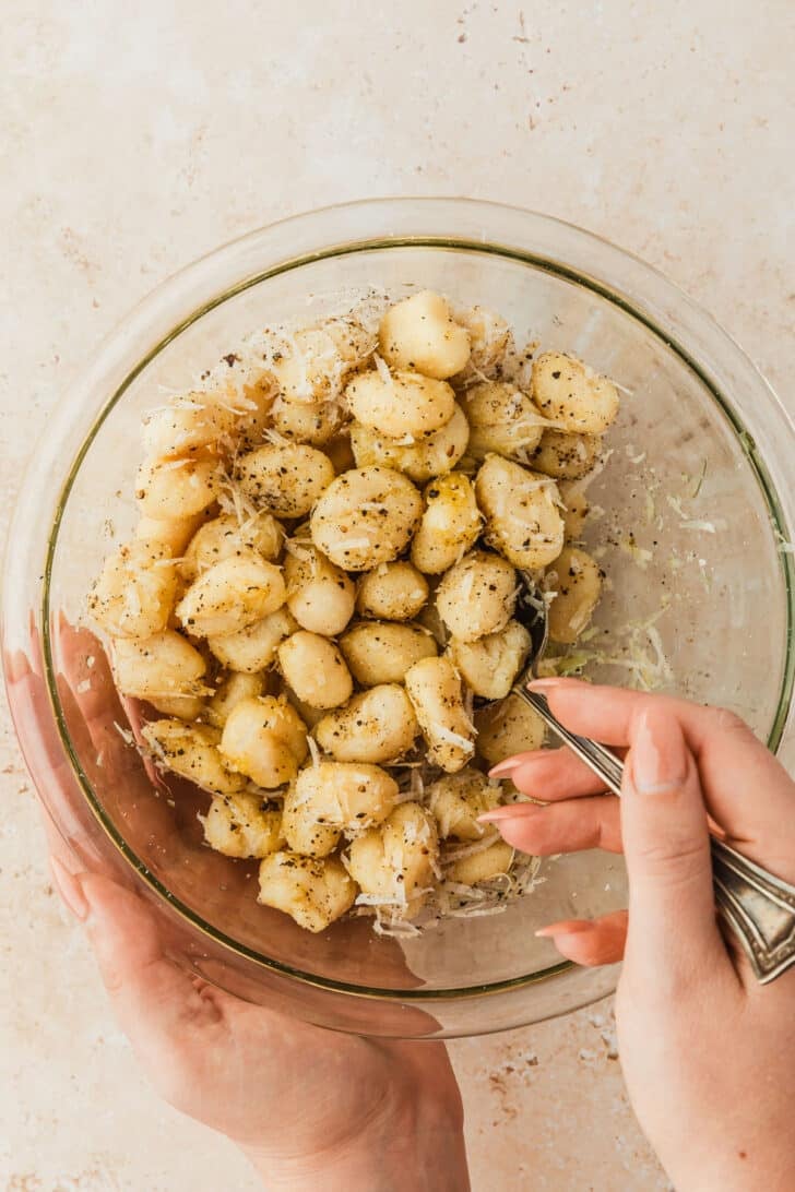 A hand using a spoon to stir gnocchi and cheese in a clear bowl.