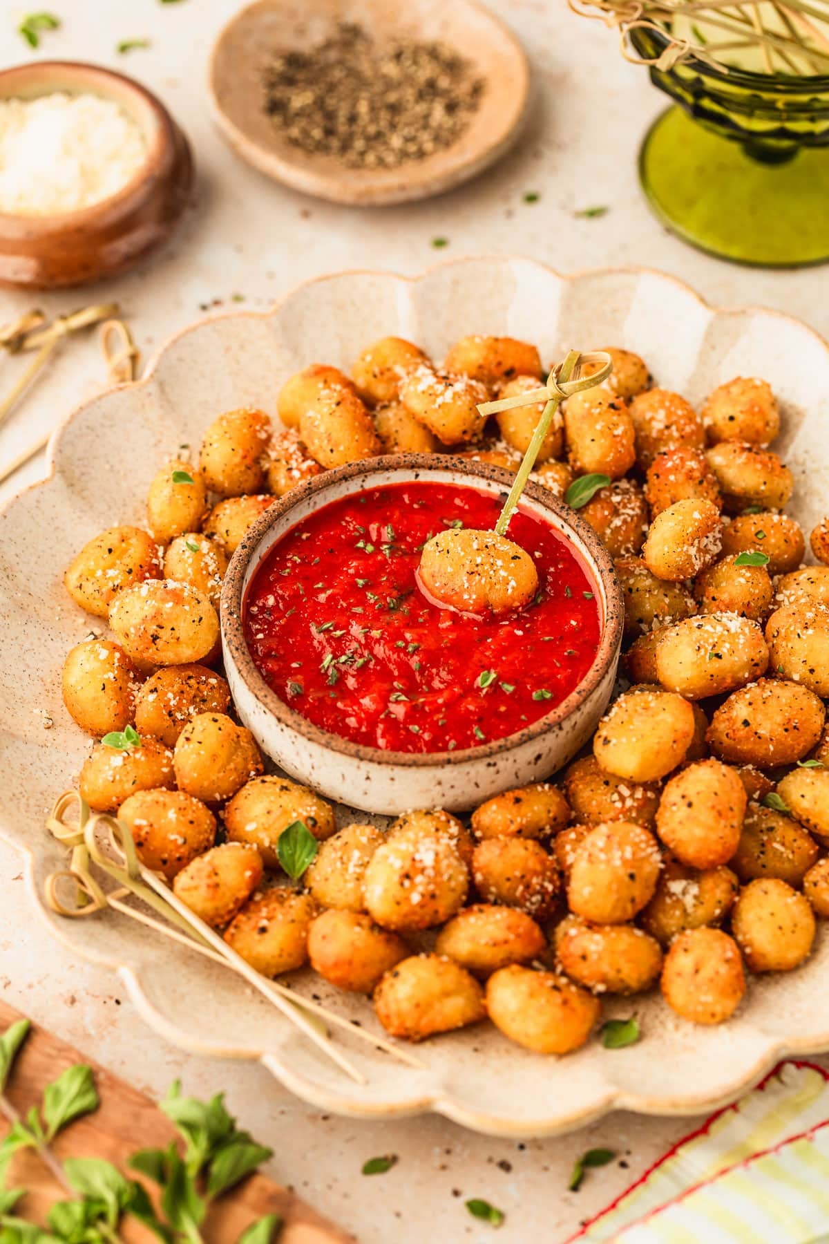 A beige platter of cacio e pepe air fryer gnocchi and a white bowl of marinara sauce on a beige counter next to a wood board of oregano, green bowl of picks, and brown bowls of pepper and cheese.