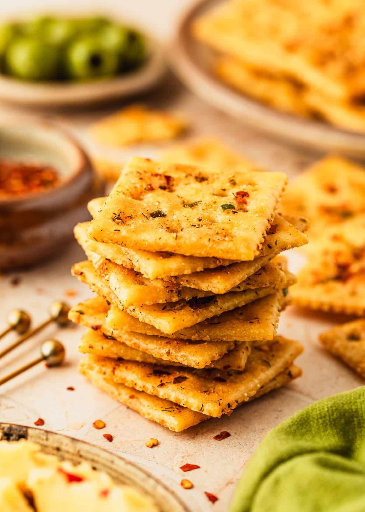 A stack of baked fire crackers next to an olive green linen and brown bowls of crackers, pepper Jack cheese, olives, and spices on a tan tiled table.