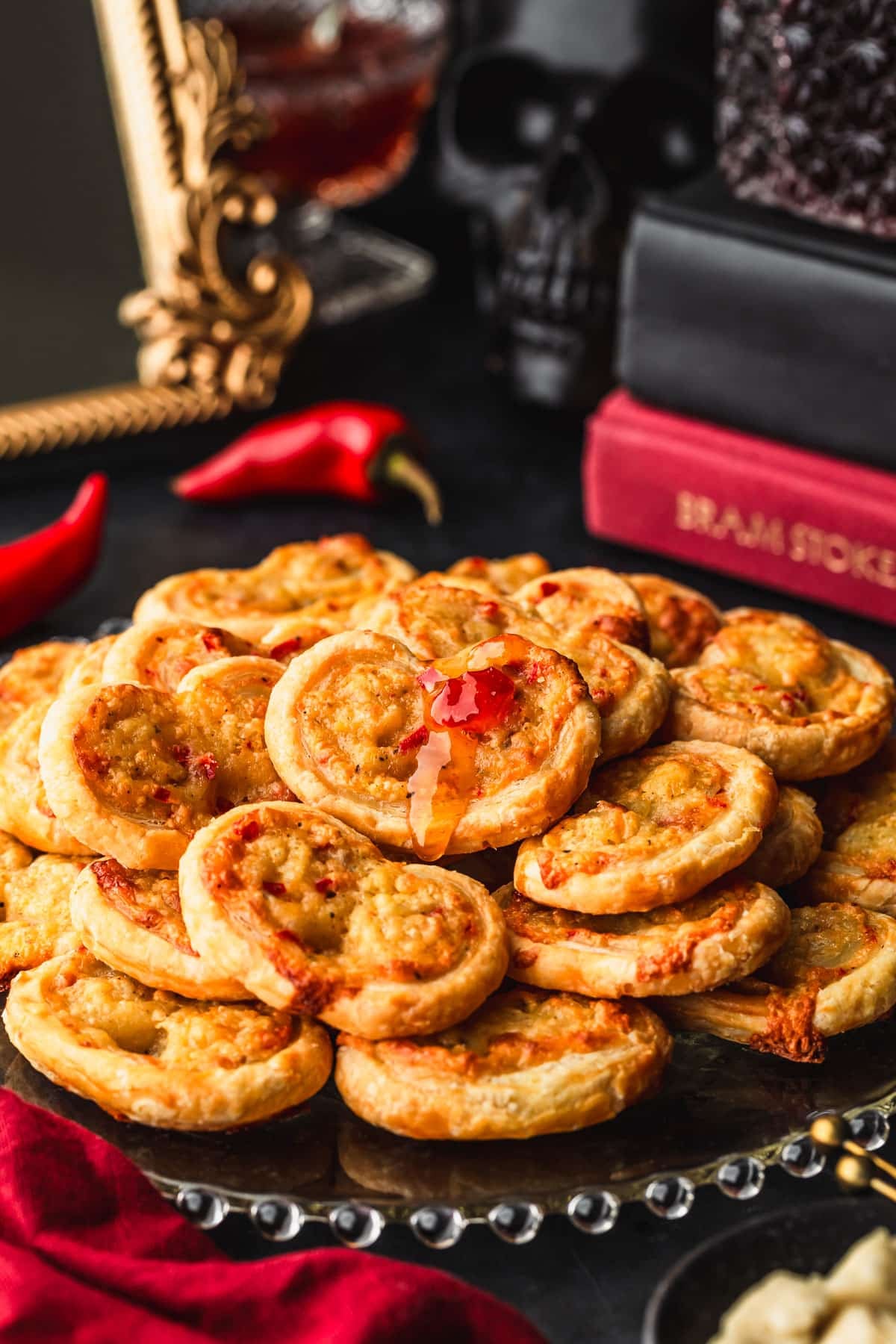 A glass platter of vampire slayer garlic cheddar palmiers on a black counter next to a black skull, a stack of books, a dark red linen, and a black bowl of cheddar cheese.