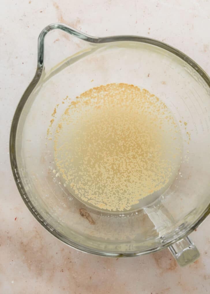 A clear mixing bowl with water, yeast, and sugar on a tan counter.
