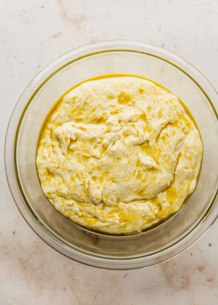 A clear bowl of dough covered in olive oil for focaccia muffins.