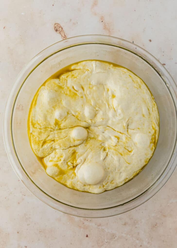 A clear bowl of fermented dough on a tan counter.