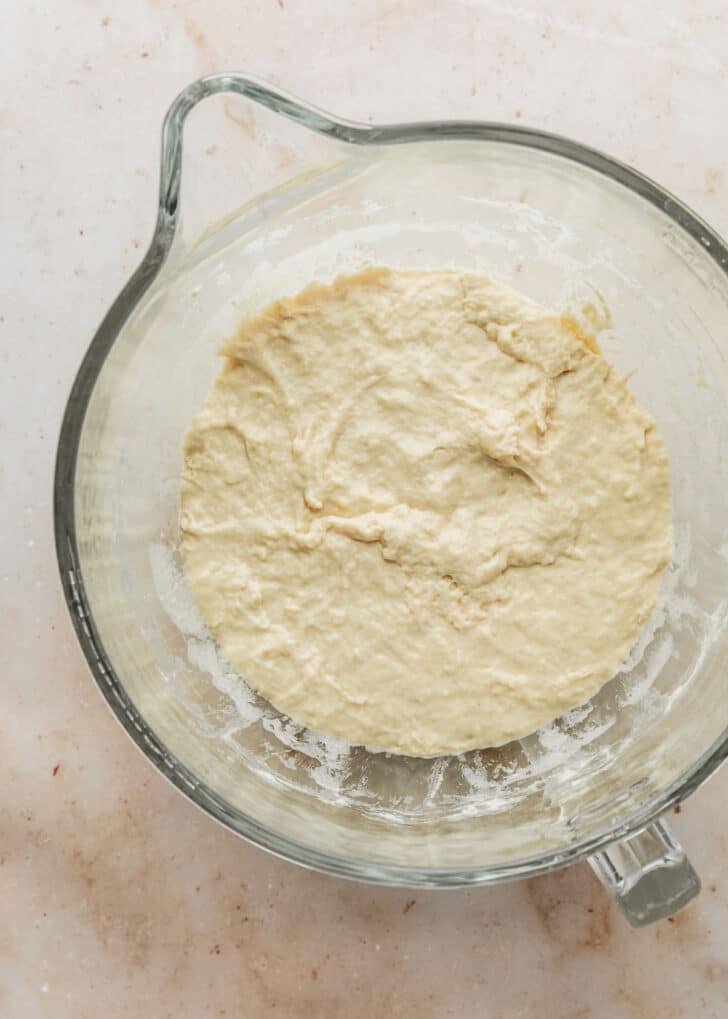 A clear bowl of dough for focaccia muffins on a brown stone counter.