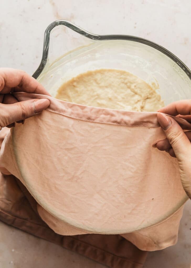Hands covering a bowl of focaccia dough with a pink linen.