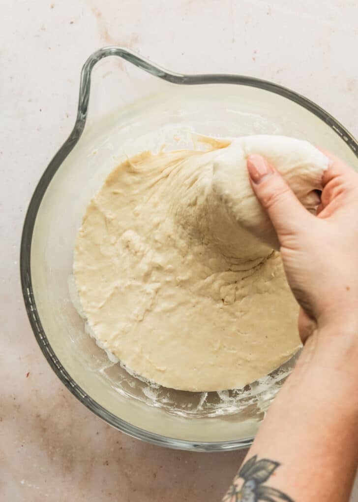 A hand stretching dough from a clear bowl.