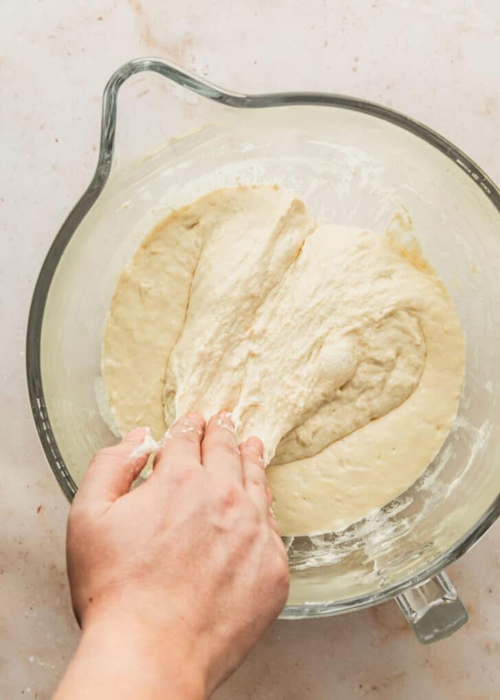 A hand folding dough from a clear bowl.