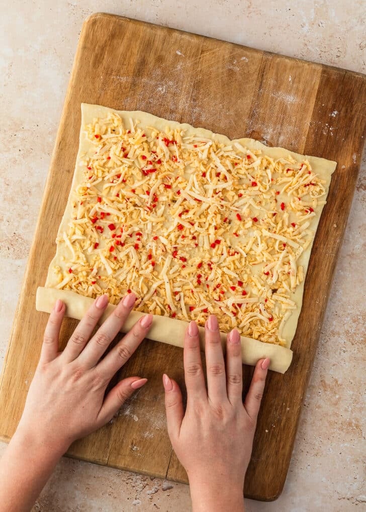 Hands rolling puff pastry covered in cheese.