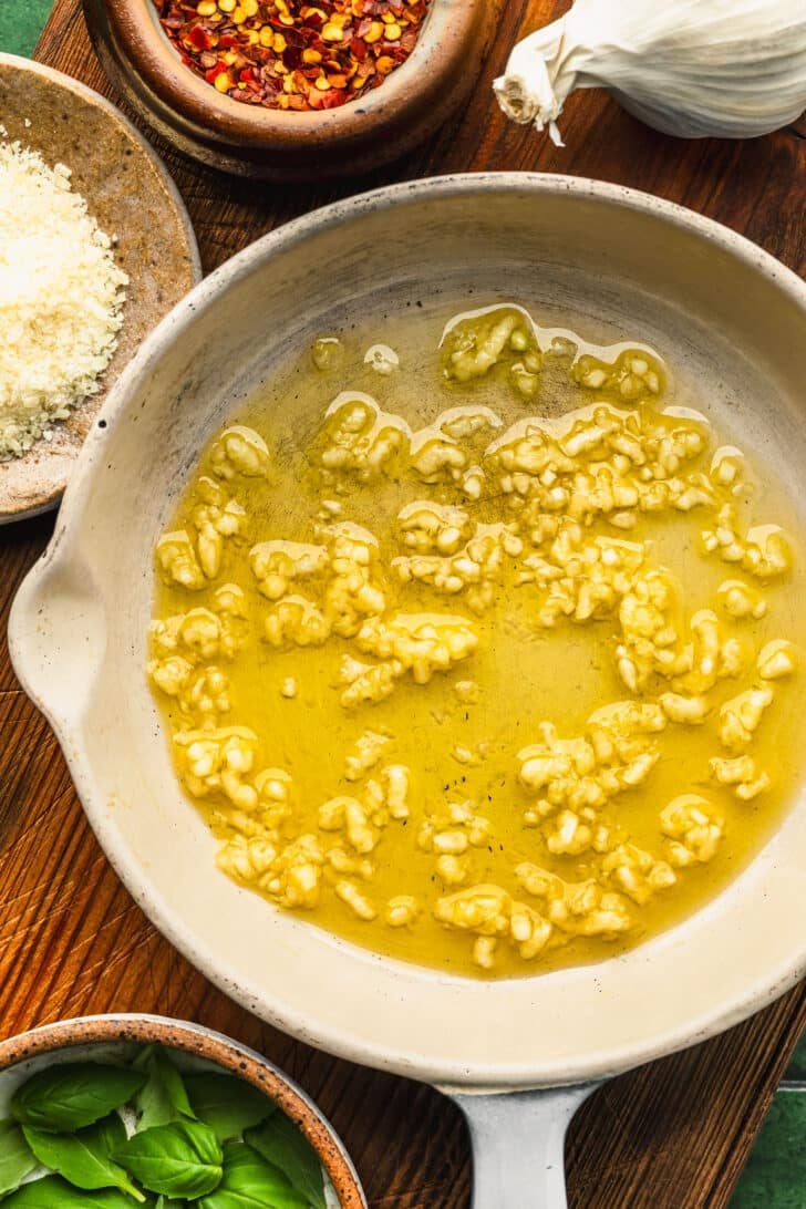 A white pan of olive oil and garlic on a wood board next to brown bowls of red pepper flakes, parmesan, and basil.