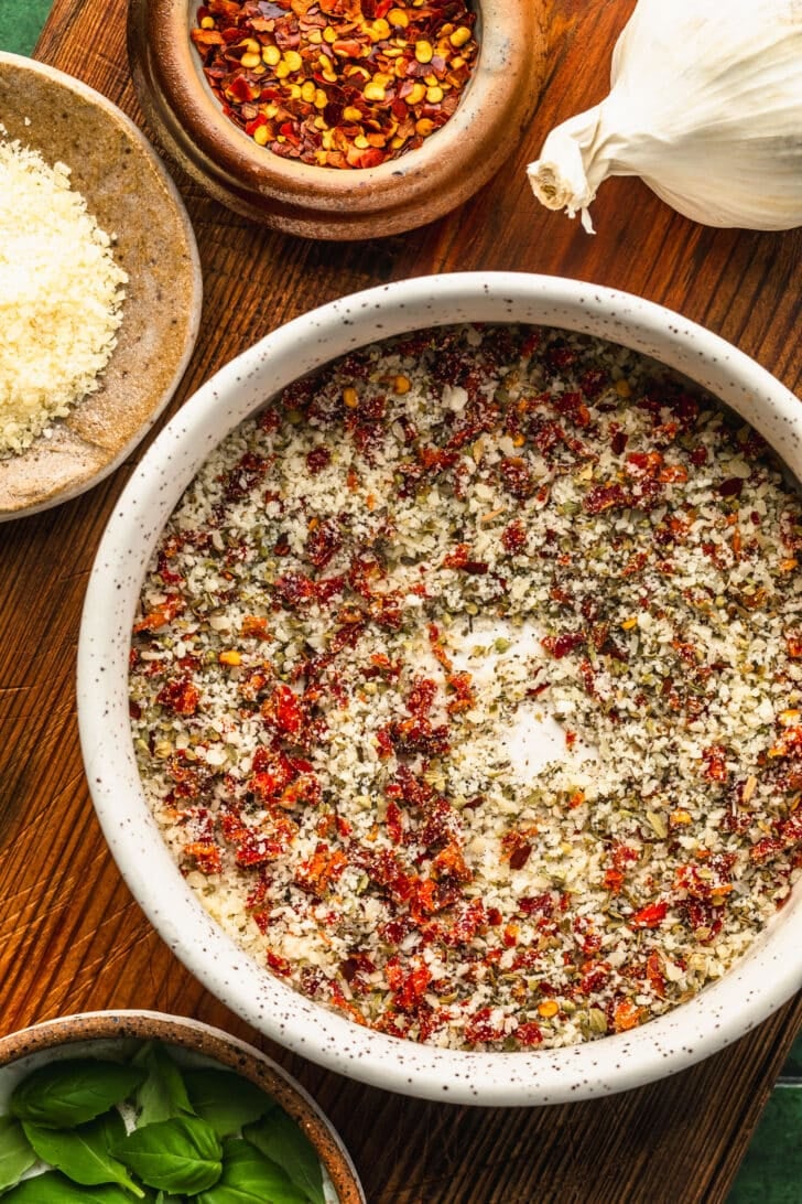 A white bowl of parmesan and herbs on a wood board next to brown bowls of red pepper flakes, parmesan, and basil.