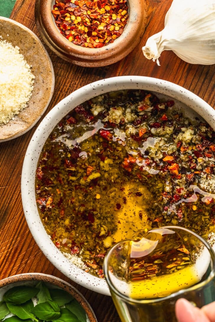 A hand pouring a jar of olive oil into a white bowl of parmesan and herbs on a wooden board next to brown bowls of red pepper flakes, parmesan, and basil.
