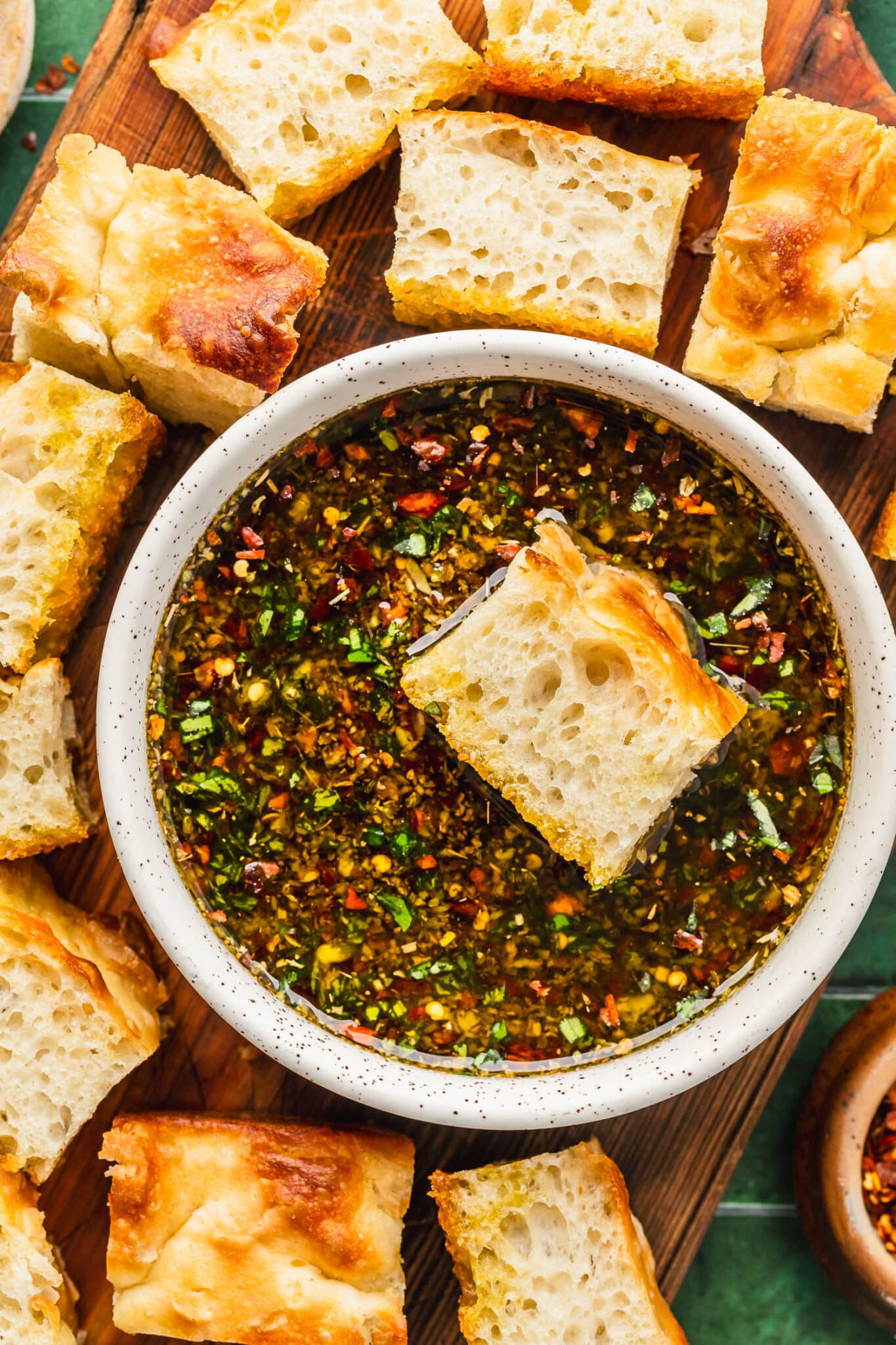 A white bowl of garlic, herb, and parmesan olive oil bread dip on a wood board of focaccia.