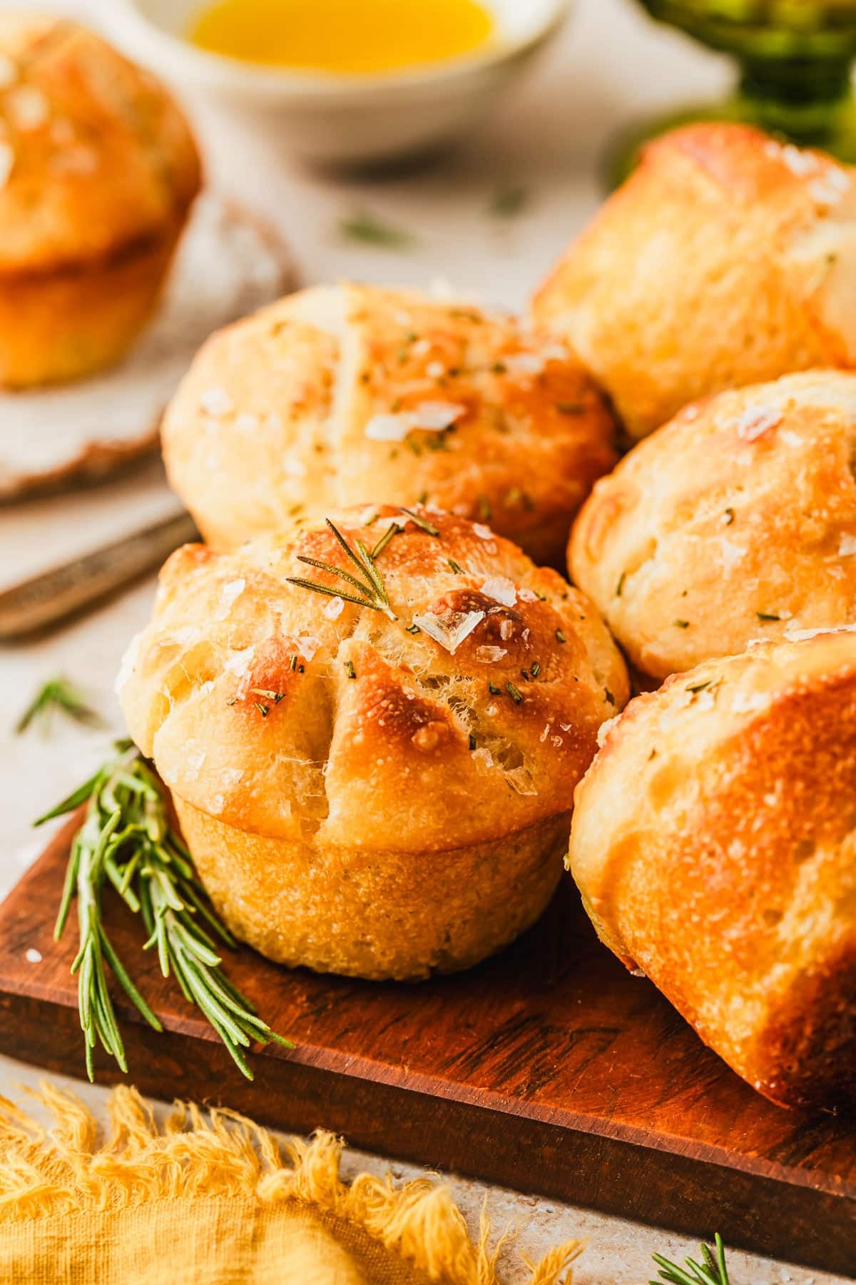 Focaccia muffins on a wooden board next to a yellow linen, a green bowl of almonds, and a white bowl of olive oil on a tan counter.