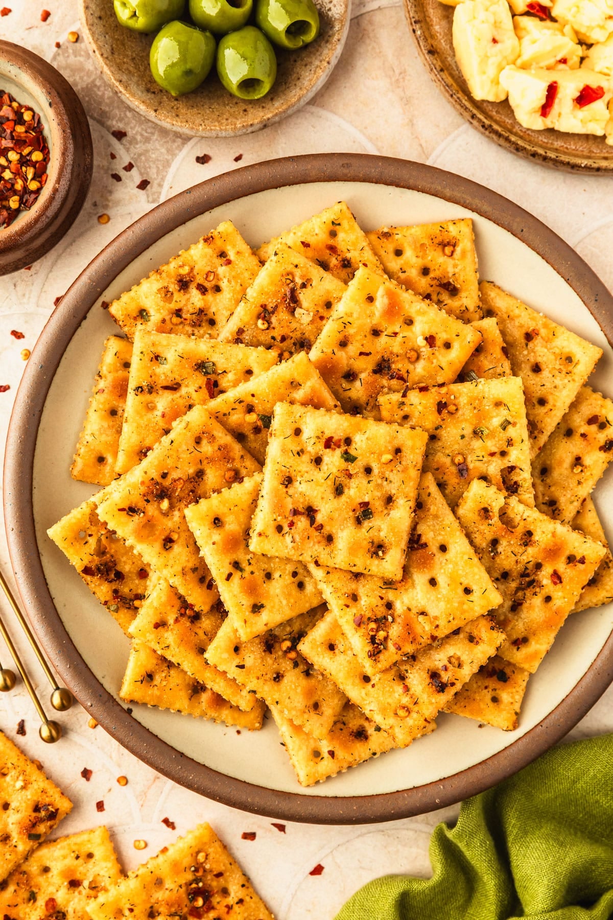A beige platter of smoky Alabama fire crackers next to brown bowls of Pepper Jack cheese, olives, and red pepper flakes and an olive green linen on a tan tiled counter.