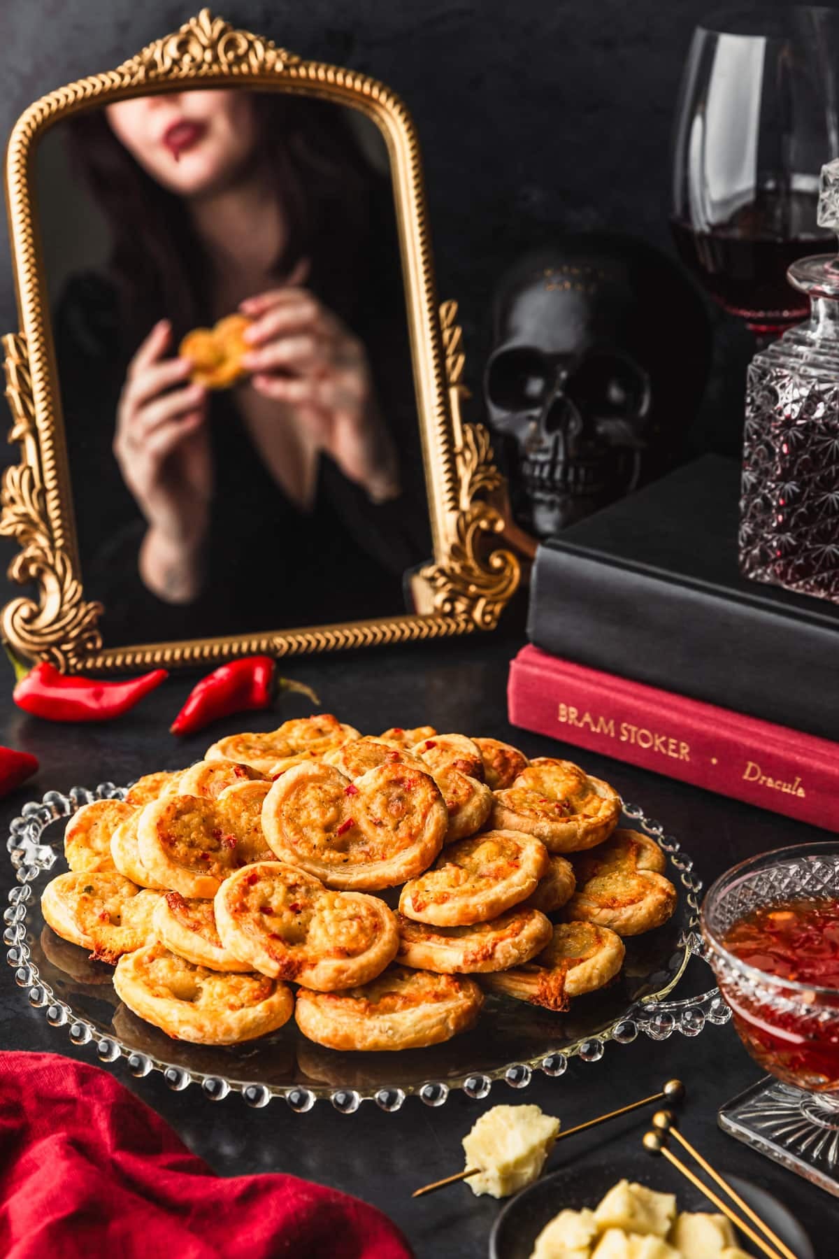 A glass platter of garlic cheddar vampire slayer palmiers on a black counter, next to stacked books, a red linen, a black skull, a black bowl of cheddar, and a gold mirror with a woman dressed as a vampire reflected in the background.