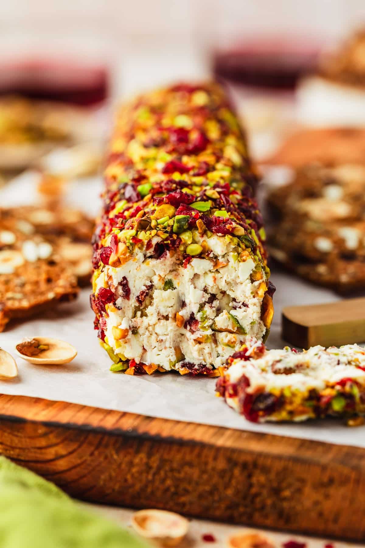 A cranberry feta cheese log with orange zest and pistachios on a wooden board next to a green linen and glasses of red wine.