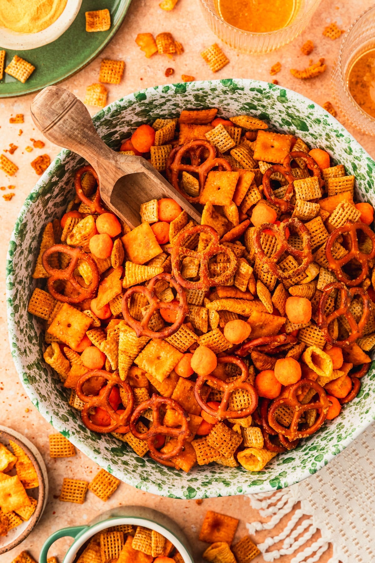 A green and white speckled bowl of homemade cheesy cheddar Chex mix on an orange counter next to glasses of beer, a green plate, and bowls of party mix.