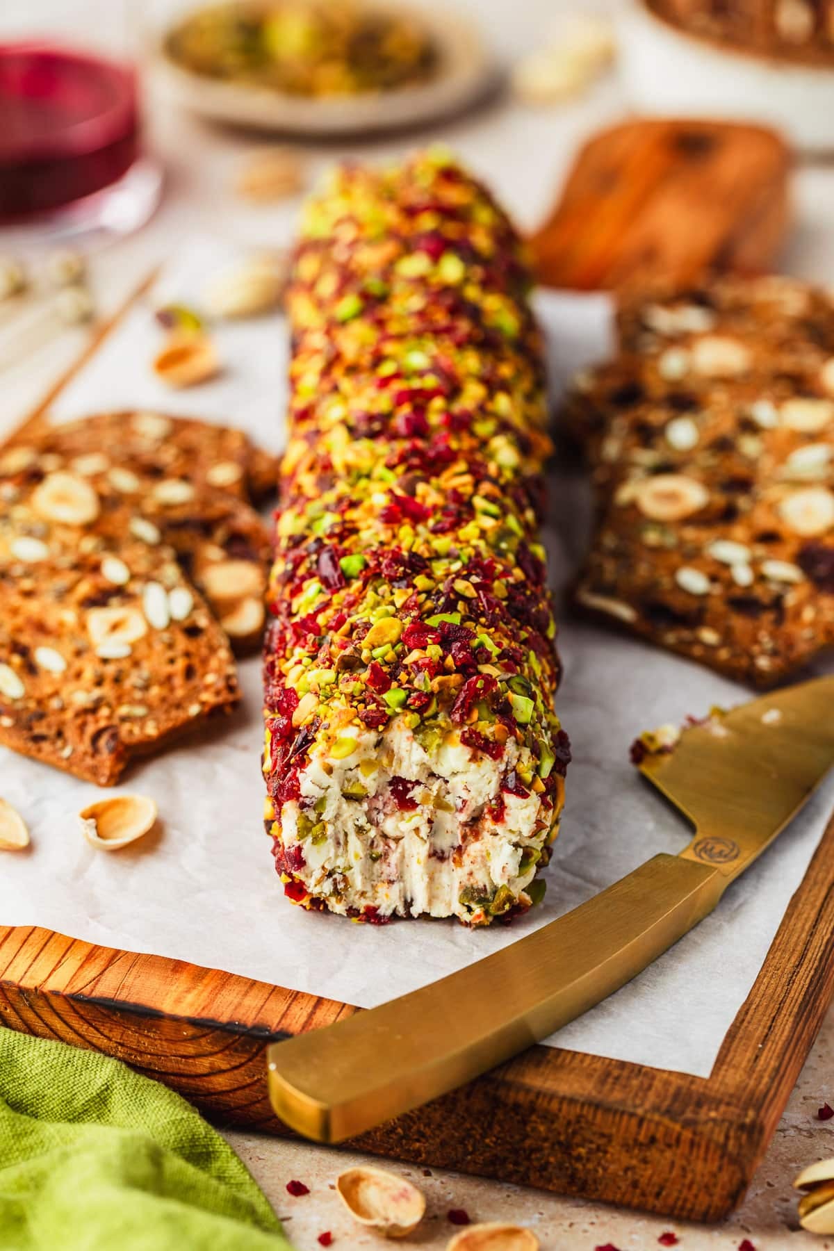 A cranberry feta cheese log with pistachios on a wood board of crackers next to glasses of red wine and a brown bowl of pistachios on a tan counter.