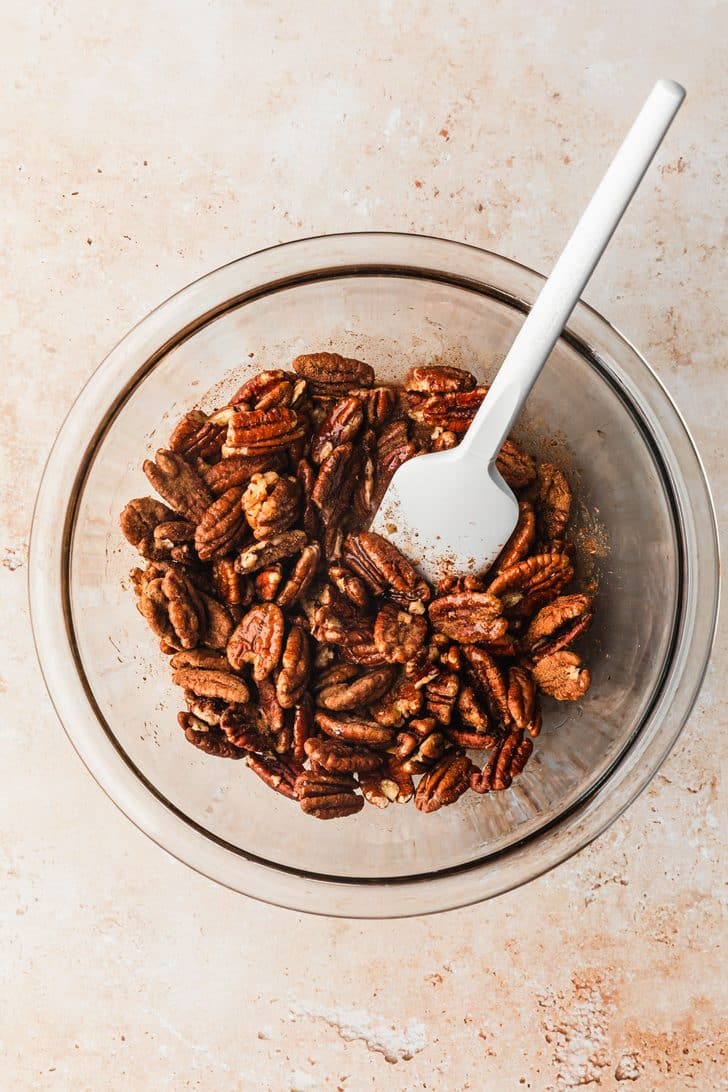 A clear bowl of pecans, maple syrup, and spices on a tan counter.