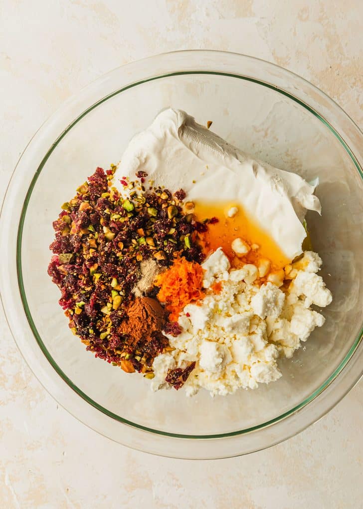 A clear bowl of cream cheese, feta, honey, chopped cranberries, chopped pistachios, orange zest, and cinnamon on a tan counter.