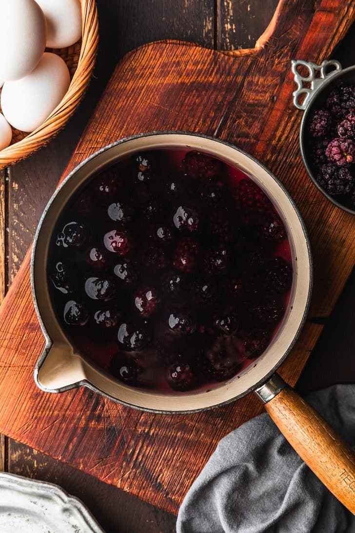A pot of blackberries and water on a wood table next to a silver bowl of blackberries, a brown bowl of eggs, and a grey linen.