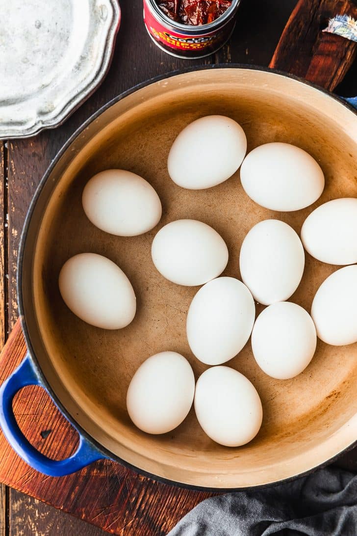 A pot of hard-boiled eggs on a wood table next to a grey linen, silver plates, and a can of chipotle peppers.