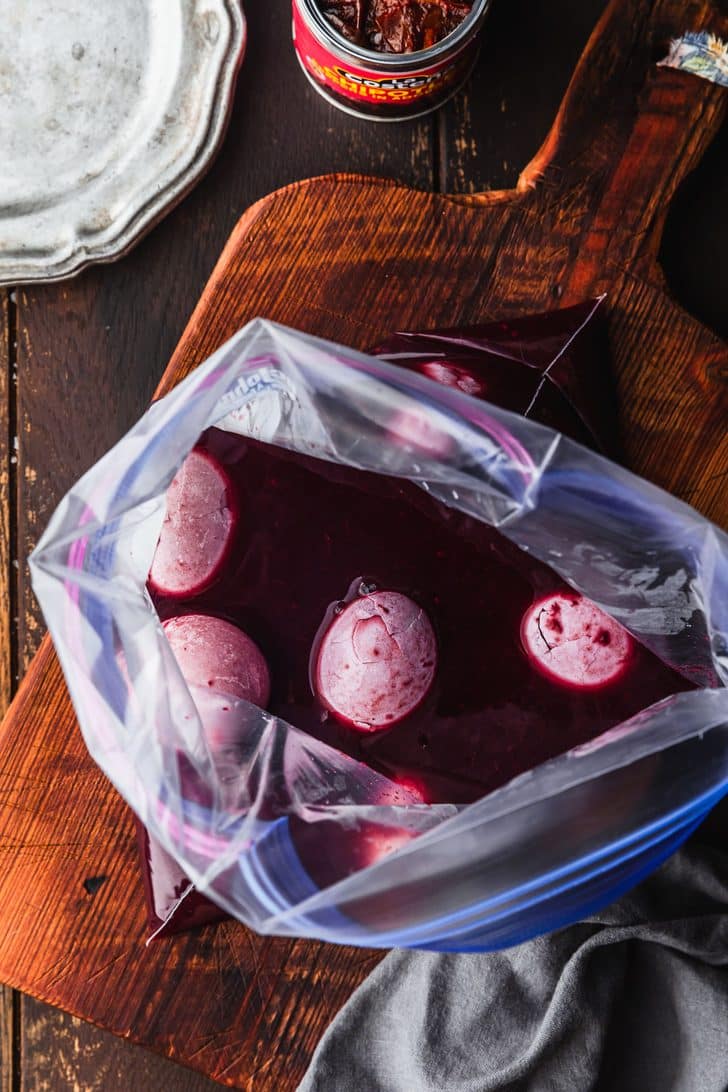 A plastic baggie of blackberry juice and hard-boiled eggs on a wood table next to a can of chipotle peppers, grey linen, and silver plates.