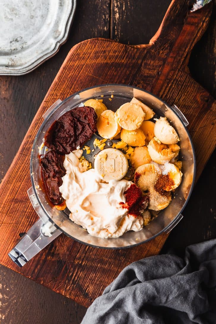 A food processor bowl filled with egg yolks, mayo, spices, and chipotle peppers on a wooden counter next to a grey linen and silver plates.