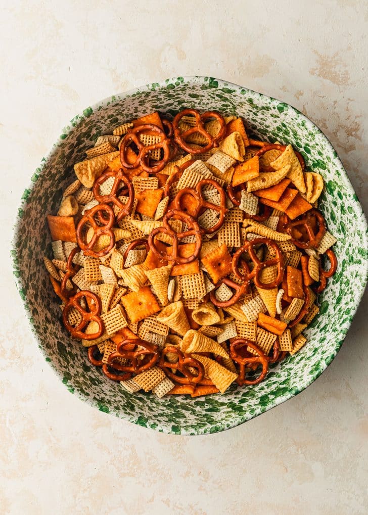 A green and white bowl of cereal, pretzels, cheese crackers, and Bugles on a tan counter.