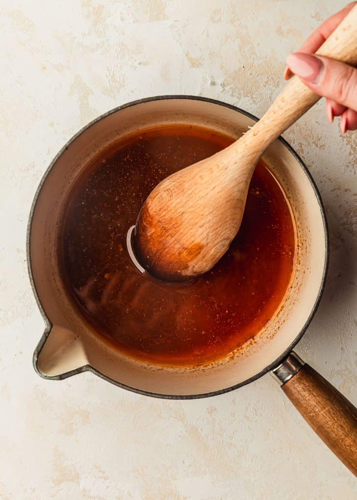 A hand using a wood spoon to stir melted butter and spices in a small saucepan.